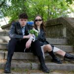Young couple in leather jackets sitting on outdoor steps holding flowers, showcasing modern style.