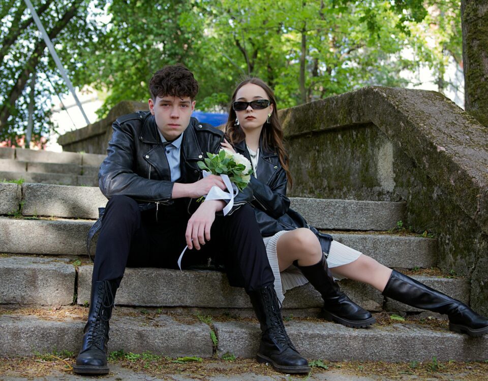 Young couple in leather jackets sitting on outdoor steps holding flowers, showcasing modern style.